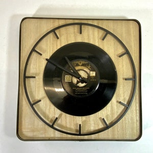 May include: A square wooden clock with a black vinyl record face. The clock has black metal hands and a black metal frame. The record has the text "Rock Around The Clock" on it.