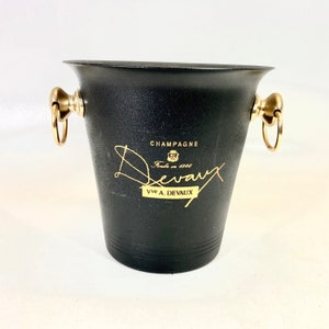 Op de afbeelding: Zwarte champagne-emmer met gouden handvatten en de tekst "Champagne De Vaux Fondée en 1818 Vive A. De Vaux" gedrukt op de zijkant.