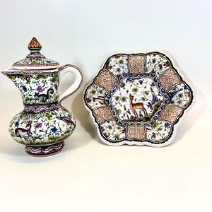 Könnte beinhalten: Eine weiße Keramikkanne mit Deckel und einem passenden Teller. Beide Teile sind mit einem Blumenmuster verziert und zeigen kleine Tierillustrationen. Die Kanne hat einen Henkel und einen Ausguss.