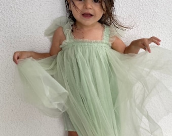 May include: A young girl wearing a light green tulle dress with ruffled straps. She is standing on a light colored floor with a white wall behind her.