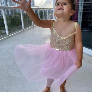 May include: A young child wearing a pink tutu dress with a gold sequined bodice and thin straps. The child is standing on a tiled surface, with one arm raised and looking upwards. The background includes a balcony railing and a view of buildings.