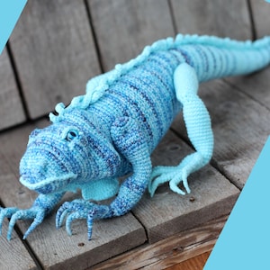 Op de afbeelding: Een handgemaakte gehaakte leguaan in blauwtinten. Het reptiel heeft een gedetailleerd ontwerp met getextureerde schubben, een lange staart en gelede ledematen. De leguaan is geplaatst op een houten oppervlak, wat zijn ingewikkelde vakmanschap en levendige kleurenpalet laat zien.