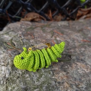 Puede incluir: Un broche de oruga de crochet verde con ojos negros. El broche está unido a un pasador de seguridad dorado.