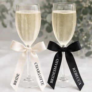 May include: Two clear glass champagne flutes filled with sparkling liquid. The left glass features a cream-colored ribbon printed with the words BRIDE and CHARLOTTE. The right glass features a black ribbon printed with BRIDESMAID and REBECCA.