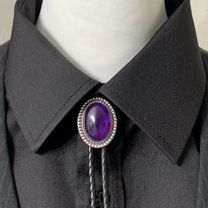 May include: A black bolo tie with a silver oval pendant featuring a purple gemstone. The tie is worn on a black collared shirt.