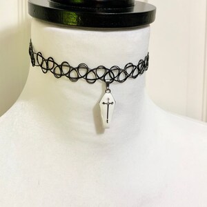 May include: A black choker necklace with a white coffin pendant featuring a silver cross. The pendant is hanging from a silver chain.