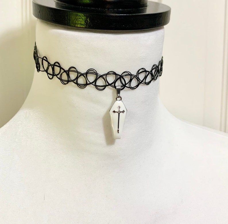May include: A black choker necklace with a white coffin pendant featuring a silver cross. The pendant is hanging from a silver chain.