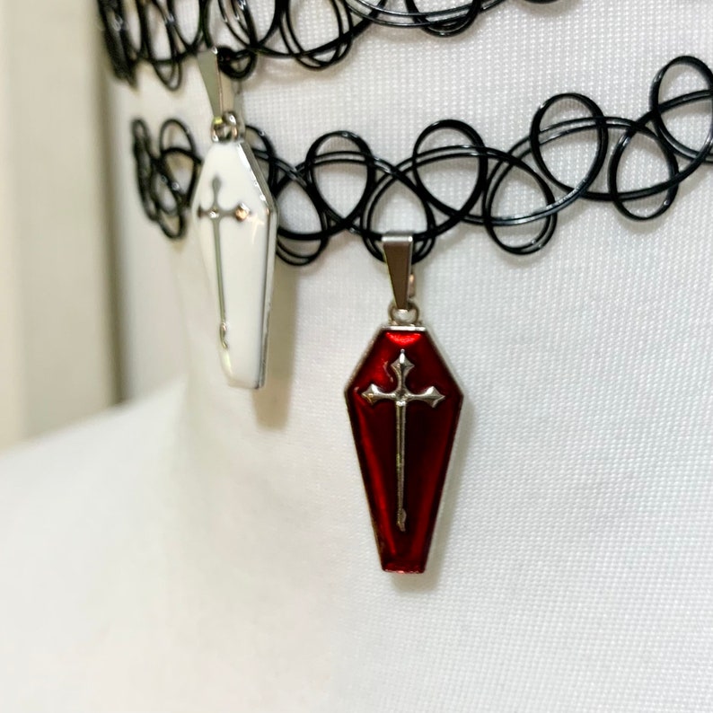 May include: Two silver coffin-shaped pendants with a cross design. One pendant is white and the other is red. The pendants are hanging from a black choker necklace.
