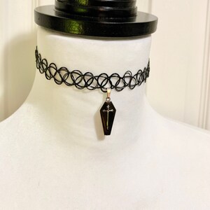 May include: A black choker necklace with a silver coffin pendant featuring a cross. The pendant is black with a gold cross.
