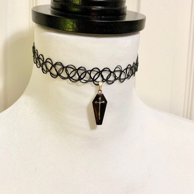 May include: A black choker necklace with a silver coffin pendant featuring a cross. The pendant is black with a gold cross.