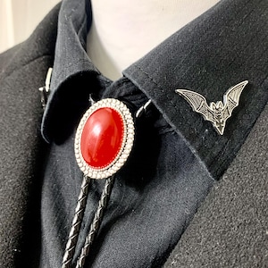 Puede incluir: Un bolo tie negro con una piedra ovalada roja y un alfiler de murciélago plateado. El bolo tie se lleva sobre una camisa negra.