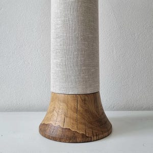 Könnte beinhalten: Eine zylindrische Lampe mit hellgrauem Leinen-Lampenschirm auf einem Naturholzsockel. Der Sockel ist ein massives, grob behauenes Holzstück, das die natürliche Maserung und Textur zeigt. Die Lampe ist für die Innenbeleuchtung konzipiert.