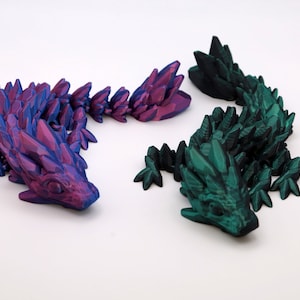 Könnte beinhalten: Zwei 3D-gedruckte Eidechsenfiguren, eine in einer tiefvioletten und blauen irisierenden Farbe und die andere in einer dunkelgrünen irisierenden Farbe. Beide Eidechsen haben eine stachelige, kristalline Textur.