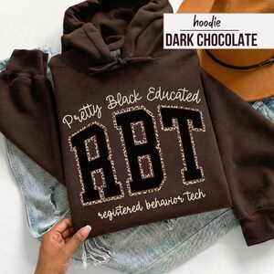 May include: Dark chocolate brown hoodie with the text "Pretty Black Educated RBT registered behavior tech" in white and black. The letters "RBT" have a leopard print pattern. A brown hat and denim jeans are in the background.