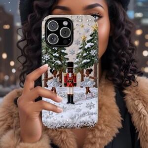 May include: A phone case featuring a winter scene with a nutcracker, ballerinas, and Christmas trees. The case has a white background with snowflakes and stars. The nutcracker is in a red uniform, and the ballerinas wear silver tutus.