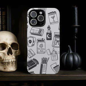 May include: A white phone case with a black and white pattern of matches, playing cards, and the text "Boo's & Booze" and "Dance with the Dead". The case is on a dark wooden shelf with a skull and a black pumpkin.