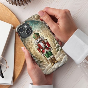 May include: A phone case with a stained glass design featuring a Nutcracker soldier in a red and green uniform. The background is a winter scene with snow-covered trees and a blue sky.