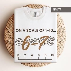 May include: White sweatshirt with the text "ON A SCALE OF 1-10..." and the numbers 6 and 7 in brown, along with corresponding emoji faces. A scale from 0 to 10 is printed at the bottom. The word "WHITE" is in the upper right corner.