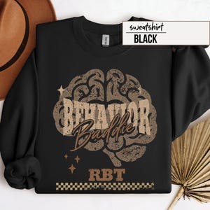 May include: Black sweatshirt with a brown brain graphic that reads "BEHAVIOR Baddie" and "RBT". The word "sweatshirt" and the color "BLACK" are in a white rectangle in the upper right corner. The design has a distressed, vintage look.