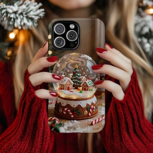 May include: A phone case featuring a snow globe design with a Christmas tree and gingerbread houses. The snow globe sits atop a decorative base with candy canes. The case is held by a person wearing a red sweater.