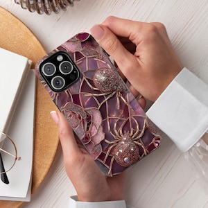 May include: A phone case featuring a design of a spider web with two spiders. The case has a purple and pink color scheme with gold accents. The spiders have a glittery texture. The case is held by a person.