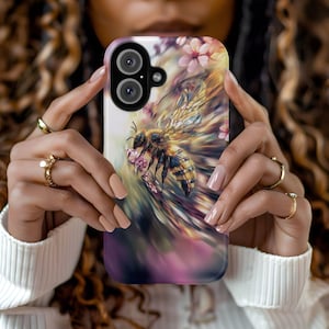 May include: A phone case featuring a detailed illustration of a bee with delicate wings, surrounded by pink flowers. The case has a gradient background of purple, yellow, and white. The phone case has a dual-lens camera.