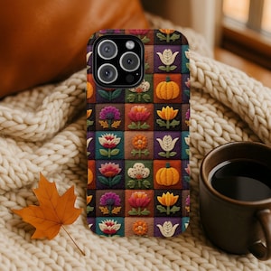 May include: A colorful phone case featuring a patchwork design with floral and pumpkin motifs. The case has a black camera module and is set against a cozy, textured background with a fall leaf and a cup of coffee.
