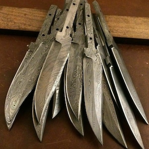 May include: A stack of 12 knife blades made of patterned steel. The blades are all the same size and shape, and they have a sharp point. The blades are arranged in a fan shape, with the points of the blades facing outward.