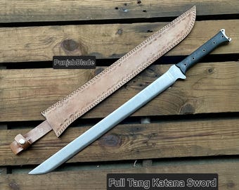 May include: A full tang katana sword with a silver blade and black handle rests beside its tan leather sheath. The sheath is decorated with a stitched border. The text "PunjabBlade" and "Full Tang Katana Sword" are visible.