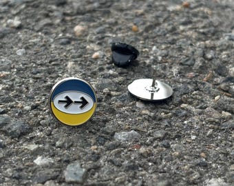 May include: A circular enamel pin featuring a horizontal design with a blue top section, a yellow bottom section, and a white center oval containing two black arrows pointing to the right. The pin includes a separate black metal clutch backing.