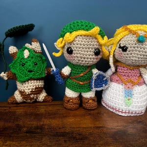 May include: Handmade crochet figures of video game characters. The figures include a green and brown Korok with a leaf, a green-clad hero with a sword and shield, and a princess in a pink and white dress. The figures have large black eyes and are made with yarn.