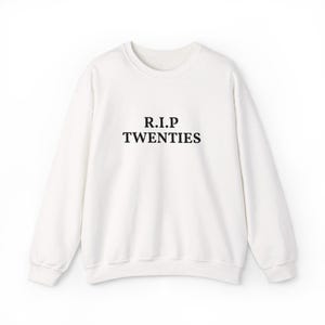 May include: A white long-sleeve crewneck sweatshirt featuring the black text R.I.P TWENTIES printed in a serif font across the center of the chest. The garment has a ribbed neckline, cuffs, and hem, with a standard fit and long sleeves.