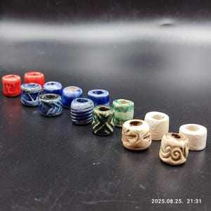 Op de afbeelding: Een verzameling keramische kralen in verschillende kleuren en ontwerpen. De kralen zijn cilindrisch met een gat in het midden. Kleuren zijn onder meer rood, blauw, groen en wit. Sommige kralen hebben een patroon, andere zijn effen.