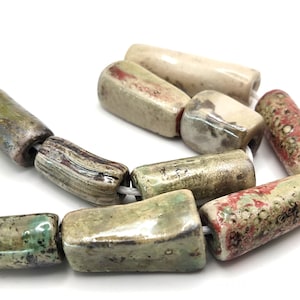 Peut inclure: Gros plan sur un fil de perles en céramique. Les perles sont rectangulaires avec des bords arrondis et une finition brillante. Les couleurs incluent le vert, le beige et le rouge. Les perles sont enfilées sur un cordon blanc.