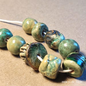 Könnte beinhalten: Eine Kette aus Keramikperlen in Grün- und Brauntönen. Die Perlen sind in verschiedenen Formen und Größen, darunter rund, oval und scheibenförmig. Einige Perlen haben eine strukturierte Oberfläche, während andere glatt sind.