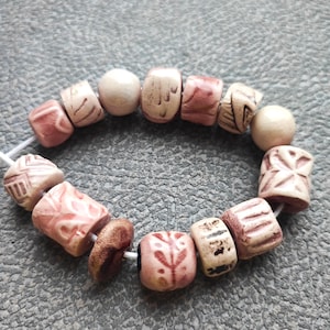 Può includere: Una disposizione circolare di perline assortite infilate su un cordino bianco. Le perline presentano forme varie, tra cui sfere e cilindri, con una tavolozza di colori nei toni del rosa tenue, crema e marrone, con motivi geometrici e floreali incisi.