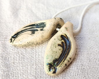 May include: Two handmade ceramic pendants with a cream base and dark blue and black decorative accents. The pendants are connected by a white cord, suggesting they are necklaces. The design features organic, flowing shapes.