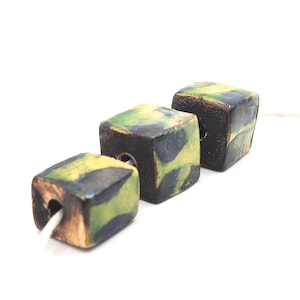 May include: Three ceramic cube beads with a green, yellow, and black glaze. The beads are strung on a white cord.