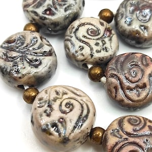 Könnte beinhalten: Nahaufnahme einer Kette aus Keramikperlen. Die Perlen sind rund, leicht abgeflacht und weisen geprägte Muster in Grau-, Braun- und Cremetönen auf. Kleine, goldfarbene Perlen trennen die größeren Perlen.