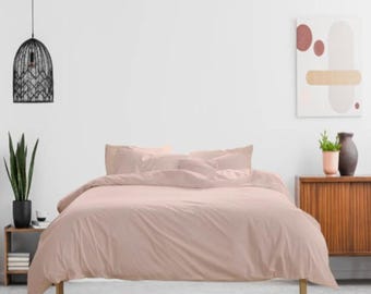 May include: A blush-colored bed set, including a duvet cover and pillowcases, is the focal point of this bedroom scene. The bedding is neatly arranged on a wooden bed frame. A black pendant light hangs above, and a potted plant sits nearby.