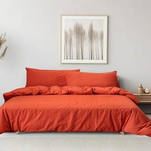 May include: A red duvet cover and two matching pillowcases on a bed. The bedding is made of a soft, textured fabric. The bed is against a white wall with a framed print of trees above the headboard.