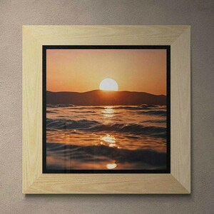 May include: A framed photograph of a sunset over the ocean. The sun is setting behind a mountain range, casting a warm glow over the water. The waves are gently lapping at the shore, creating a peaceful and serene scene.