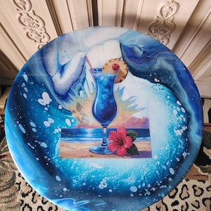 May include: Round, blue and white decorative table with a tropical cocktail design. The table features a vibrant blue cocktail with a pineapple slice and cherry, set against a beach scene. The table's edge has a wave-like pattern.