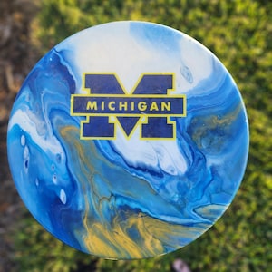 May include: A round, blue, white, and gold coaster with a blue "M" and the word "MICHIGAN" in yellow letters.