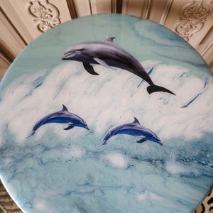 May include: Round table top with a marine theme. The table features a blue and white ocean design with three dolphins leaping from the waves. The dolphins are shades of blue and gray.
