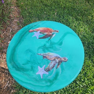 May include: A teal blue round table with a resin finish features two sea turtles and two starfish. The turtles are swimming in a swirling pattern.