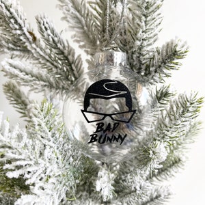 Op de afbeelding: Transparante glazen kerstbal met een zwart-witte afbeelding van Bad Bunny met een zonnebril en de tekst "Bad Bunny".
