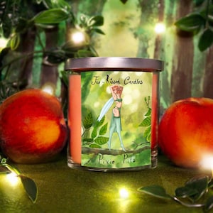 Puede incluir: Una vela de vidrio en un frasco con tapa plateada y una ilustración verde, blanca y rosa de un hada con el texto "Fairy Kissed Candles" y "Pixie Pints". La vela está rodeada de follaje verde y dos melocotones.