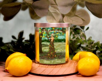 May include: A glass candle jar with a gold lid and a label that reads "The Kissed Candles, Queen of Tarts". The label features an illustration of a woman standing in a lemon tree. The candle jar is sitting on a wooden surface with three lemons in front of it.