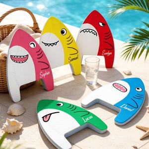 May include: Five shark-shaped kickboards in pink, yellow, red, green, and blue, each featuring a white belly, a cartoon face, and a personalized name: Sophia, Samuel, Evelyn, Lucas, and Daniel. The boards have a textured surface and fins.